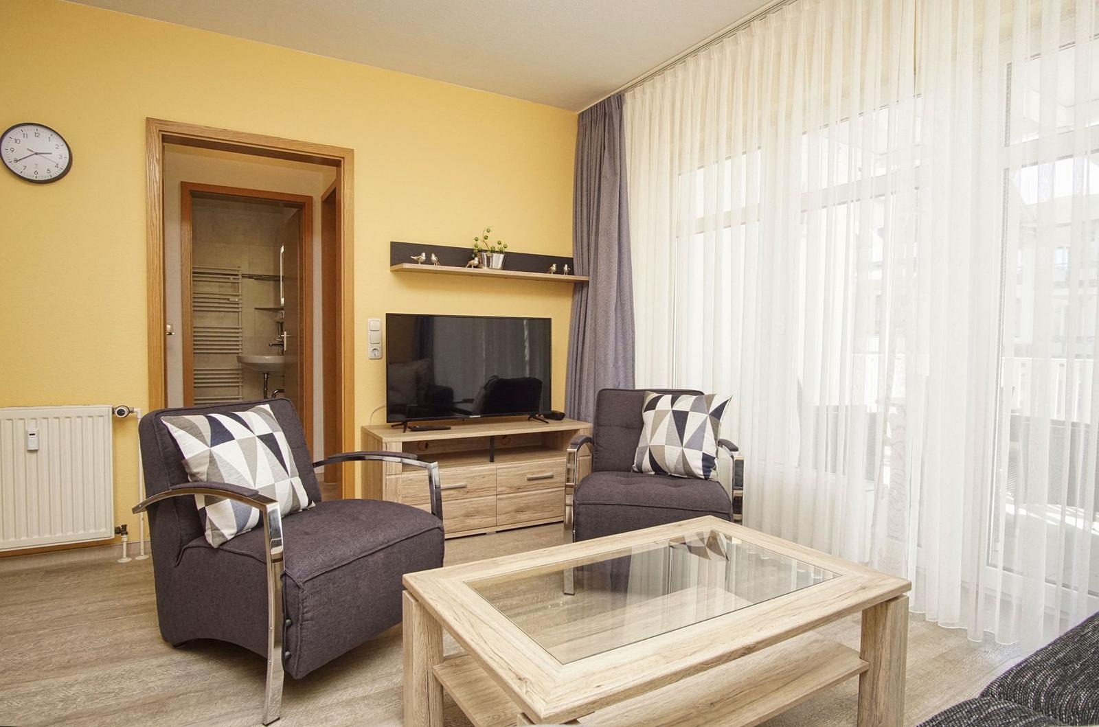 Wohnzimmer mit zwei Sesseln, Glas-Tisch und Fernseher. Hintergrund: Bad und Fenster mit Vorhängen.