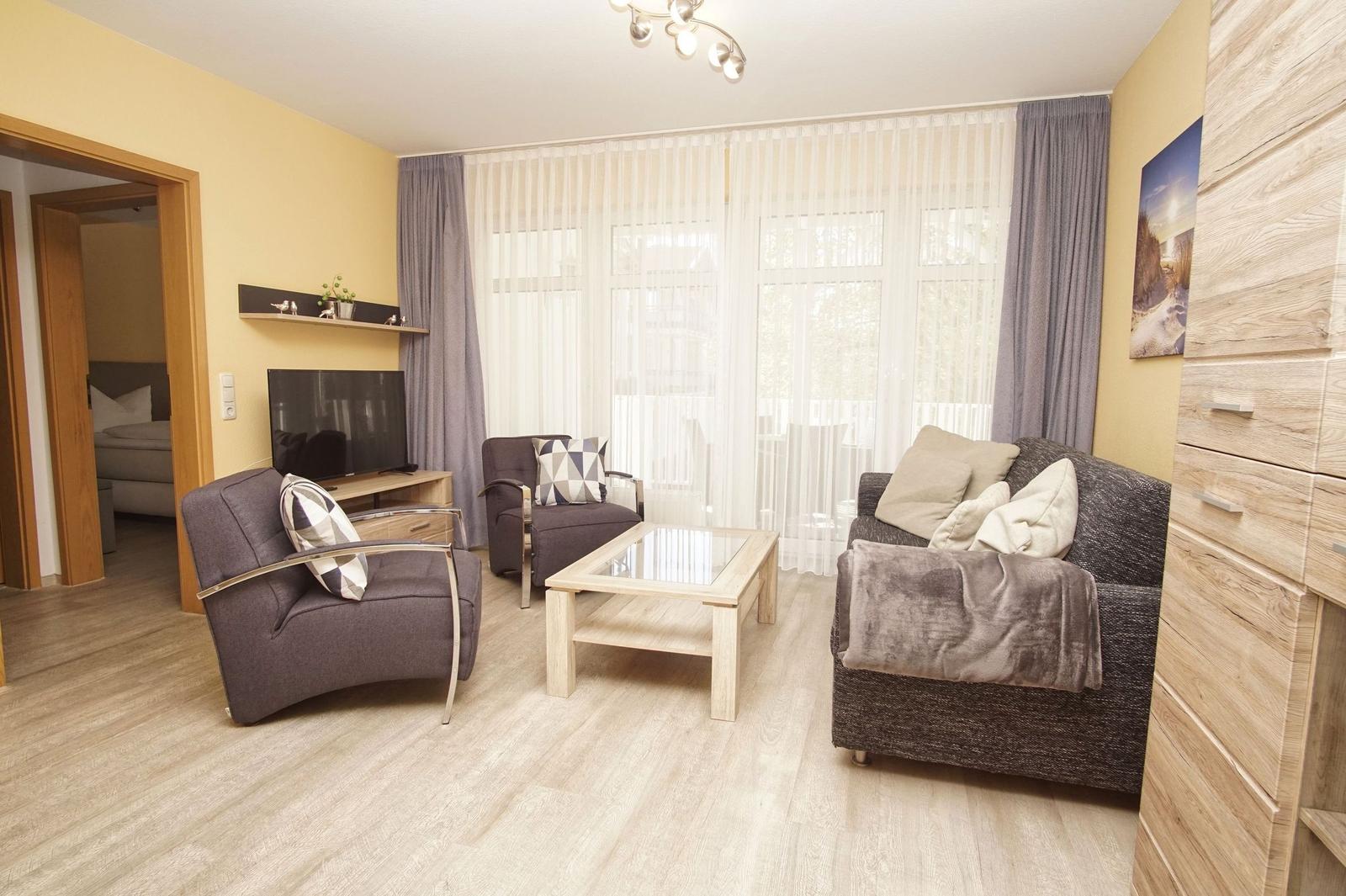 Wohnzimmer mit Sofa, zwei Sesseln, Glas-Tisch und TV. Durchgang zum Schlafzimmer.
