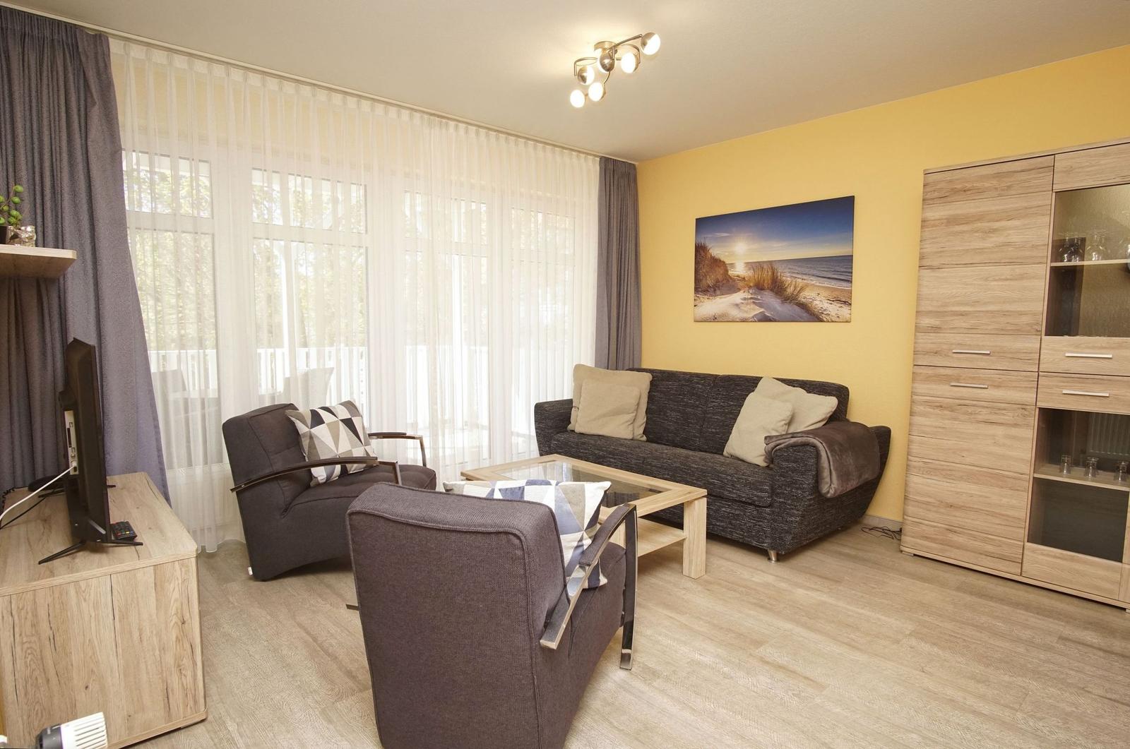 Wohnzimmer mit Sofa, Sesseln, Glas-Tisch und Fernseher. Gelber Wandbereich mit Bild.