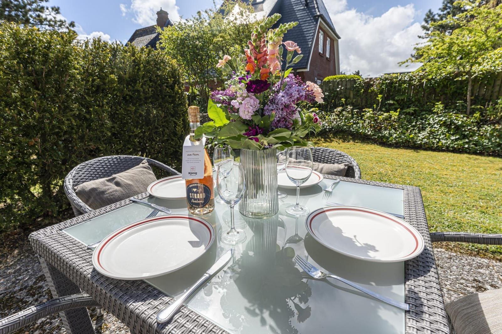 Garten-Tisch mit Blumen, Gläsern und Platzsetzung. Hintergrund: Grünfläche und Haus.