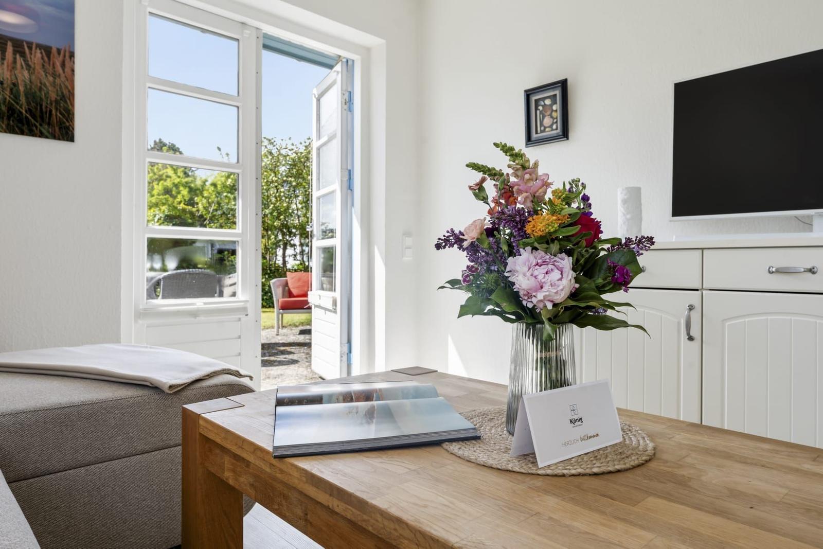 Ein Wohnzimmer mit Holztisch, Blumen und TV. Durch die Tür ist ein Garten sichtbar.