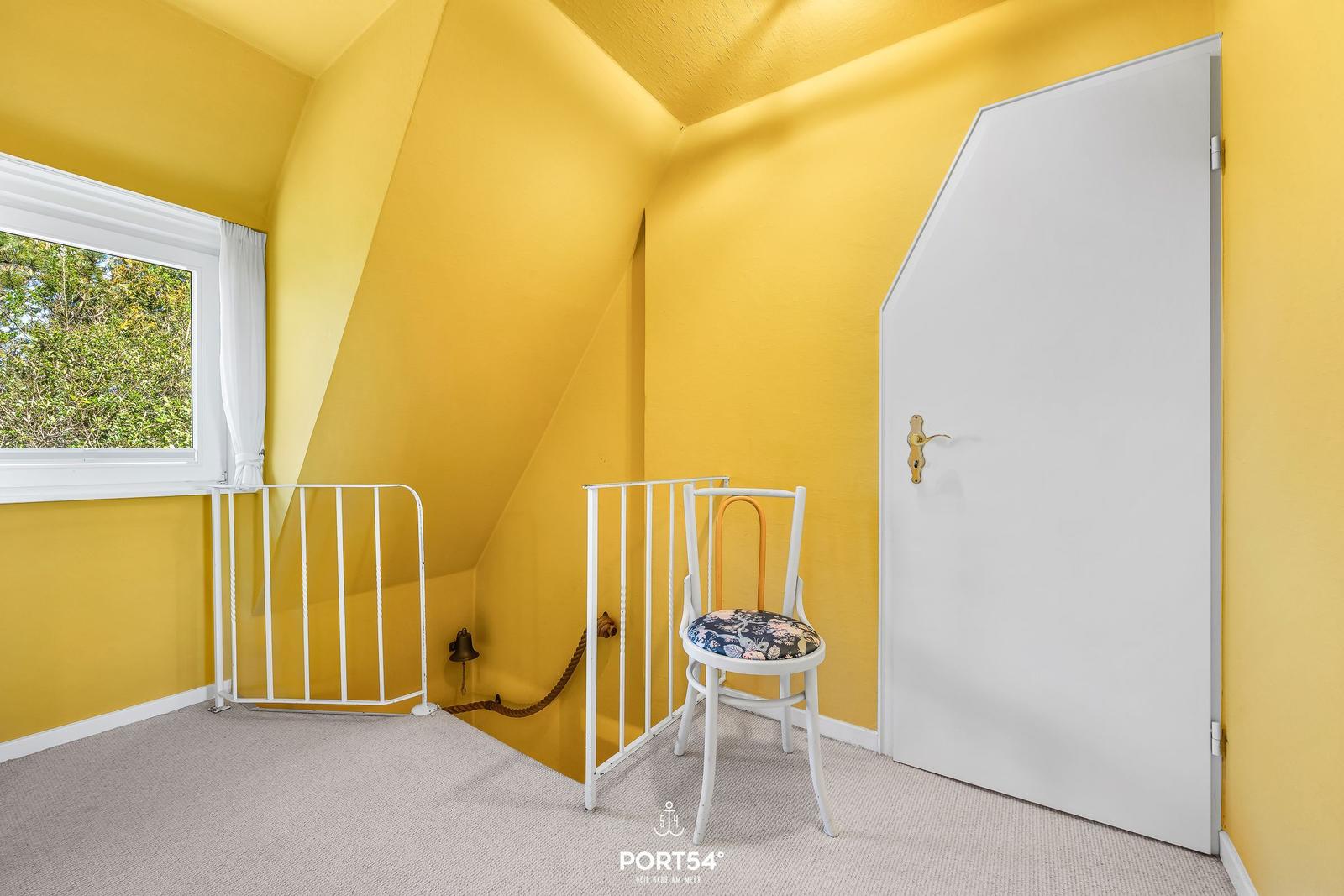 Zimmer mit gelben Wänden, Fenster, Tür und Stuhl mit floraler Sitzfläche.