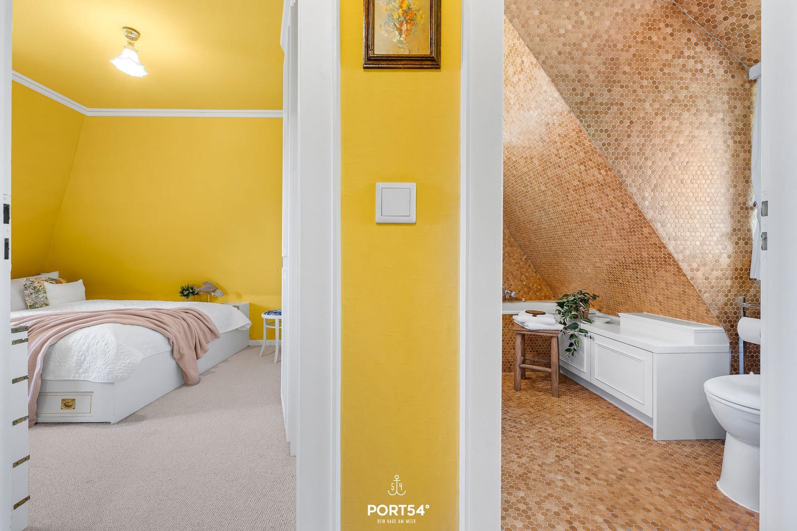 Schlafzimmer mit gelben Wänden und weißem Bett. Neben dem Bad mit Mosaikwand.