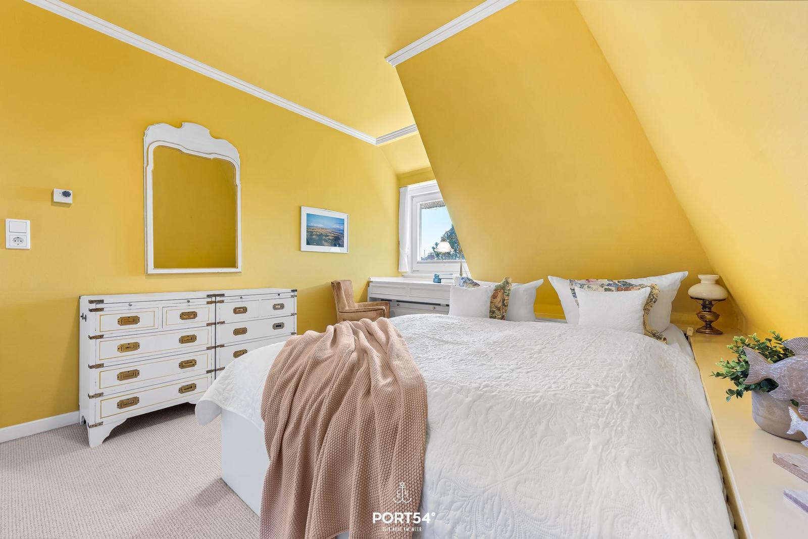 Schlafzimmer mit gelben Wänden, weißem Bett und Schrank. Fenster mit Blick nach draußen.