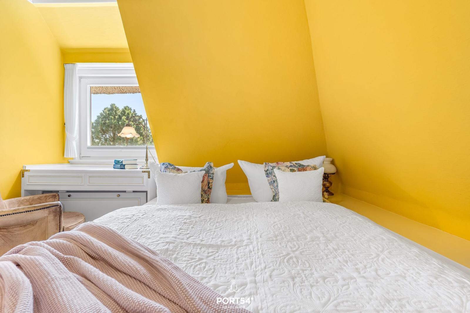Schlafzimmer mit gelben Wänden, weißem Bett und Fenster mit Blick auf Bäume.