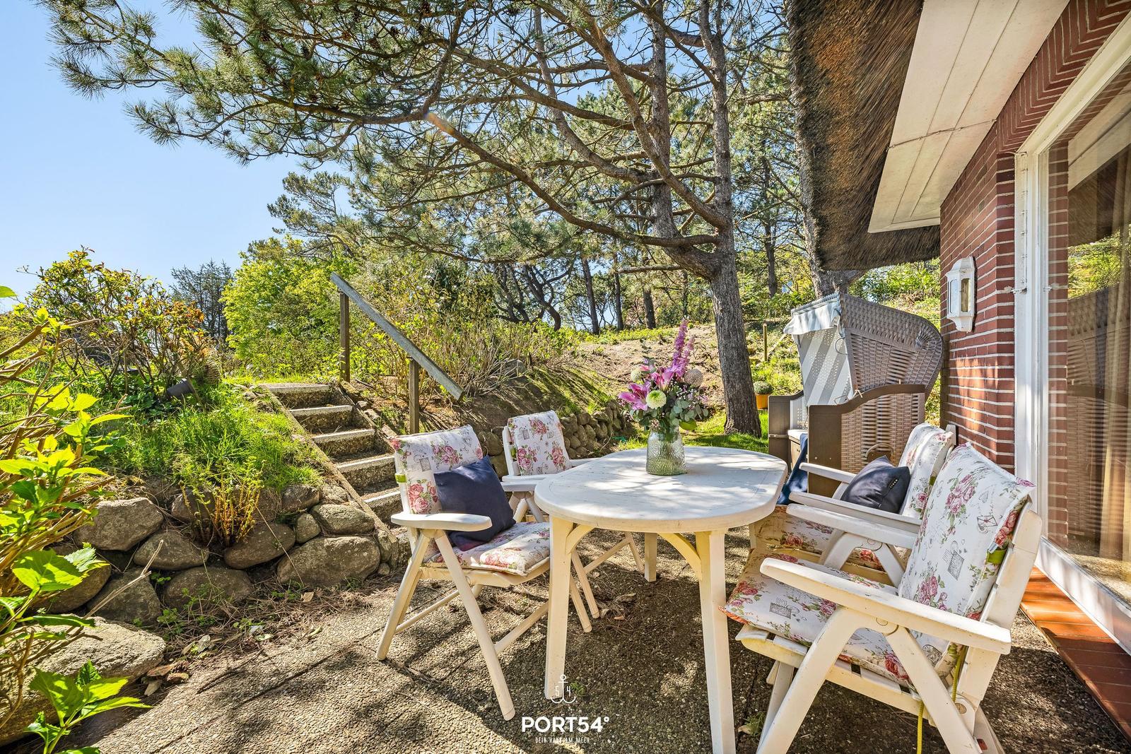 Außenbereich mit Tisch, Stühlen und Blumen unter Bäumen neben einem Haus.