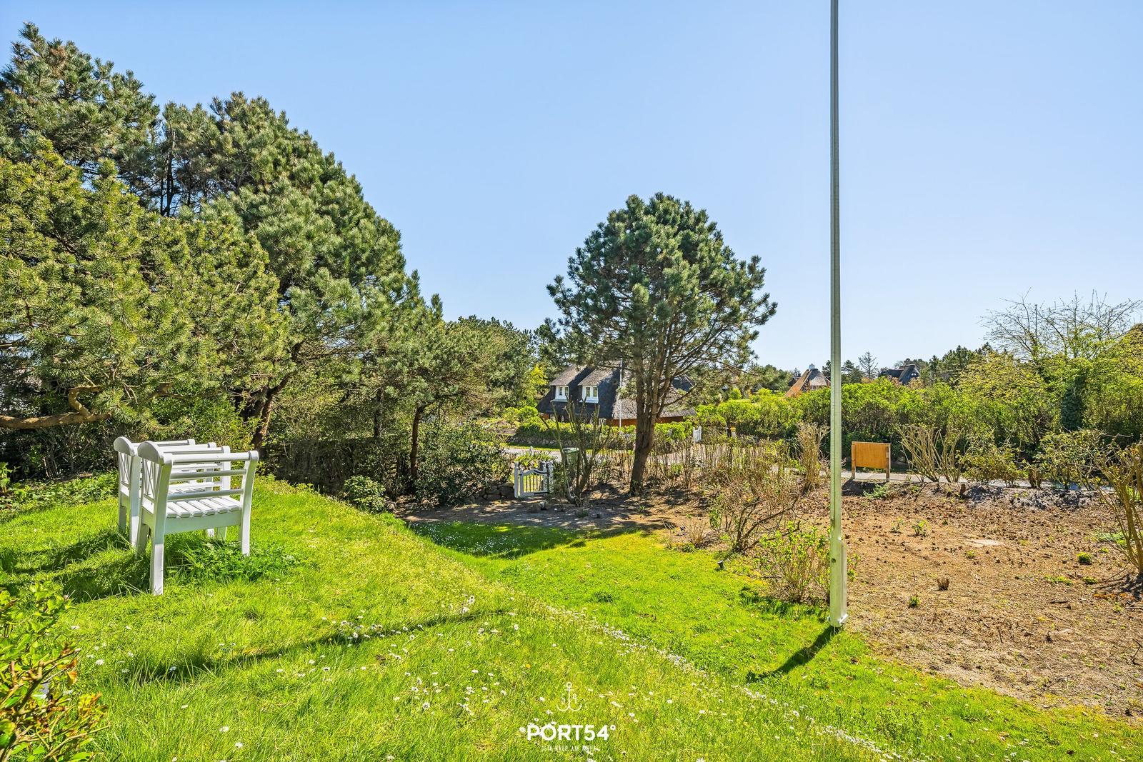 Grüner Garten mit weißem Holzstuhl, Bäumen und Blick auf Ferienhaus im Hintergrund.