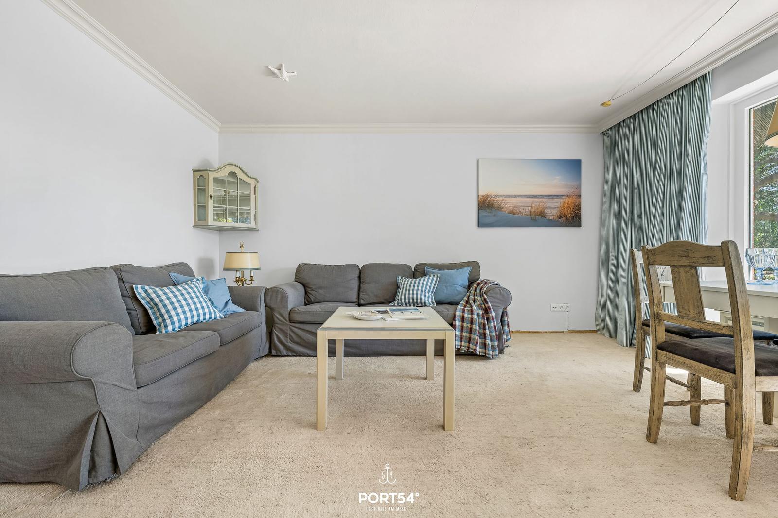 Wohnzimmer mit zwei grauen Sofas, Tisch und Stühlen. Wandbild und Gardinen sind sichtbar.