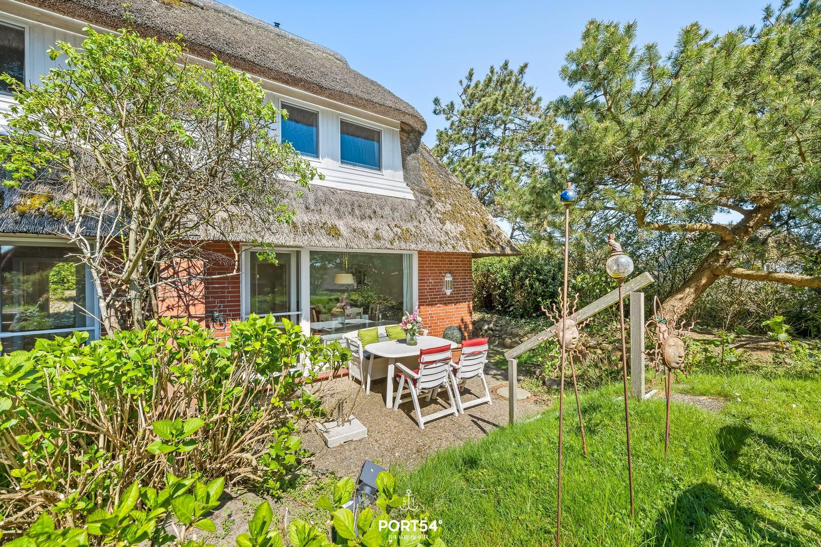 Haus mit Strohdach, Terrasse mit Tisch und Stühlen, umgeben von Grün und Bäumen.
