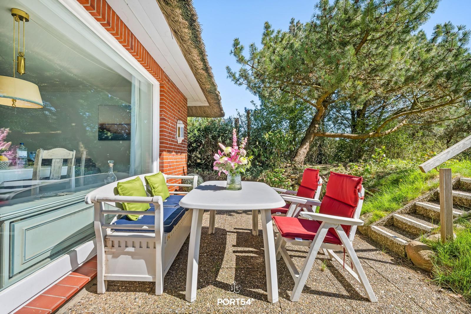 Terrasse mit weißem Tisch, zwei roten Stühlen und einer Bank mit grünen Kissen.