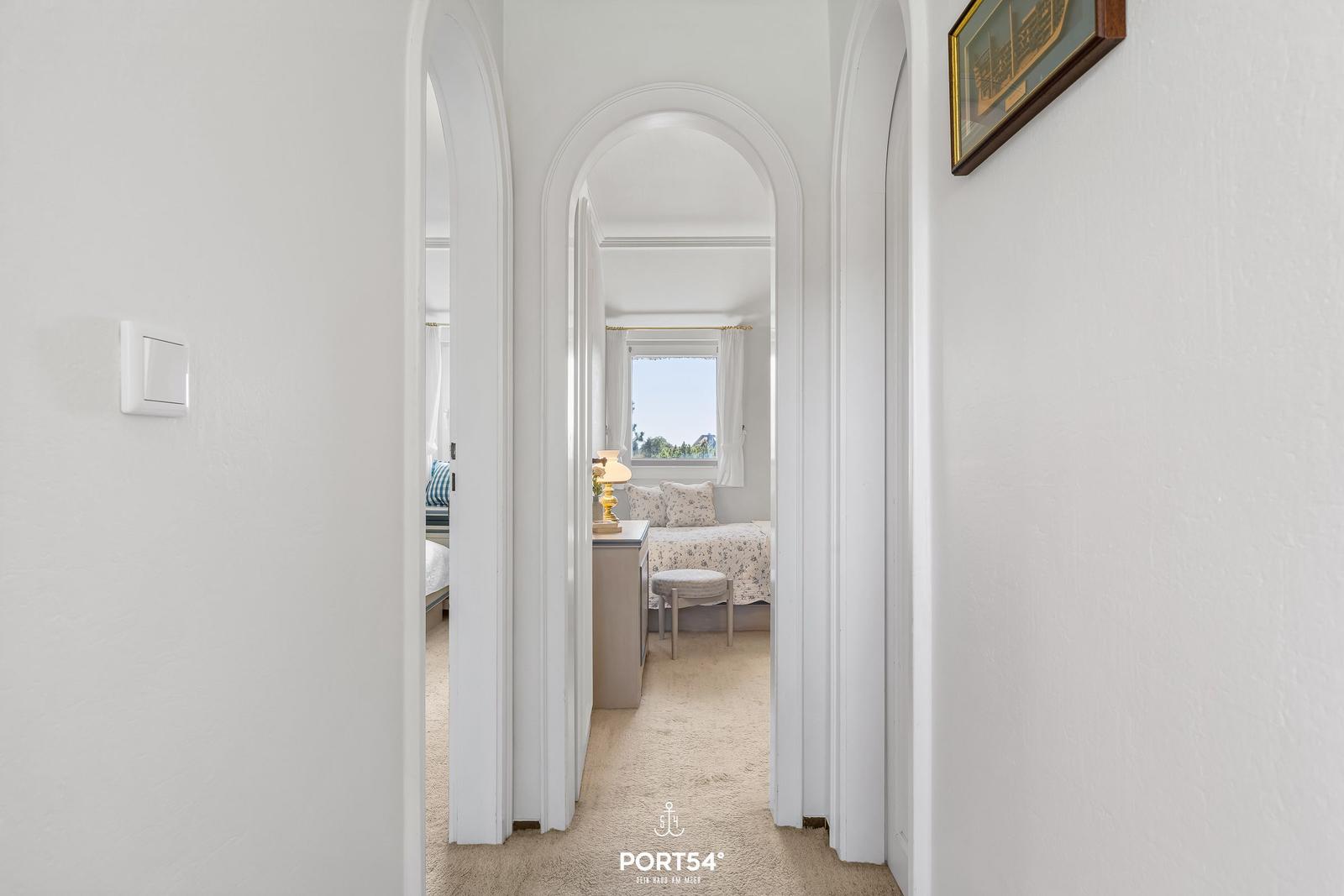 Durch einen Flur mit Bögen ist ein Schlafzimmer mit Fenster und Bett sichtbar.