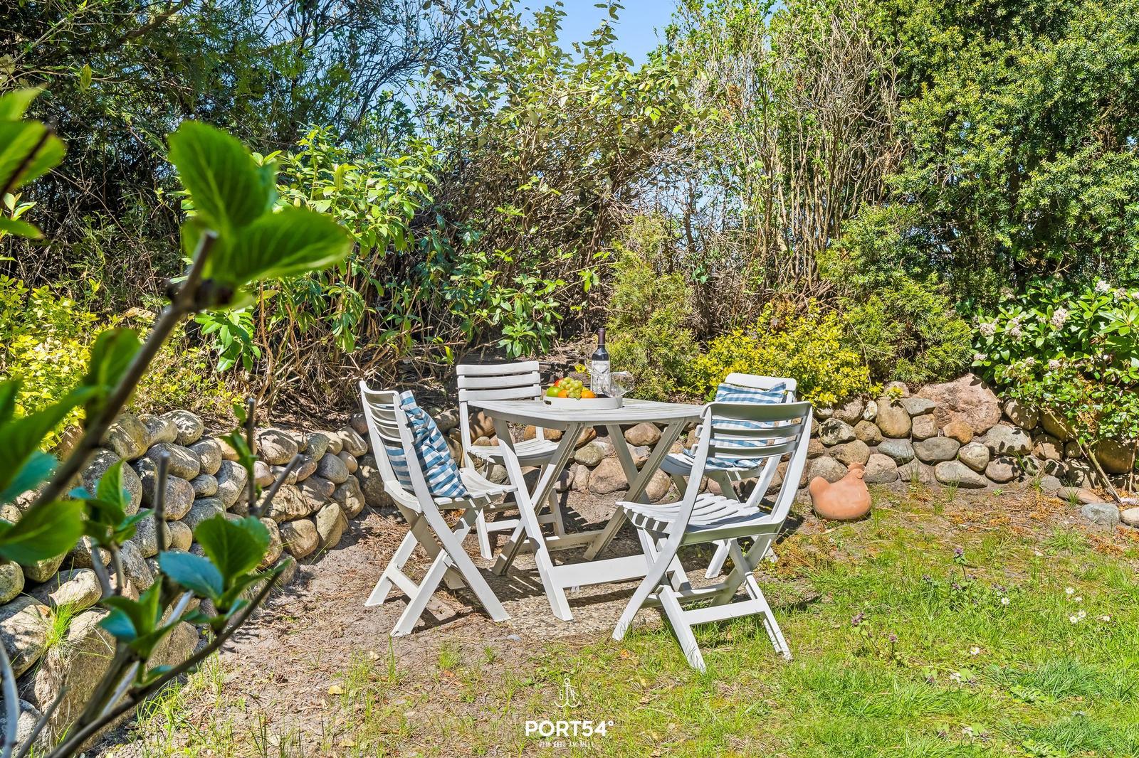 Weiße Gartenmöbel um einen Tisch mit Wein und Obst. Steinmauer und grüne Pflanzen im Hintergrund.