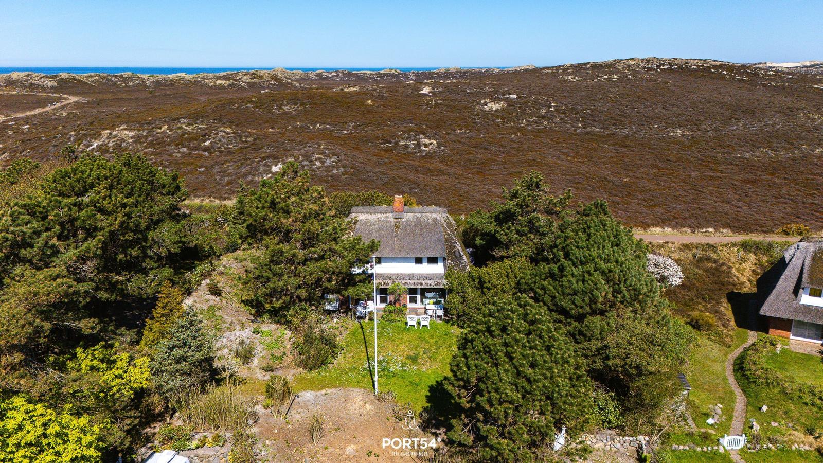 Haus mit Dach aus Stroh, umgeben von Bäumen und Grünflächen, Blick auf das Meer.