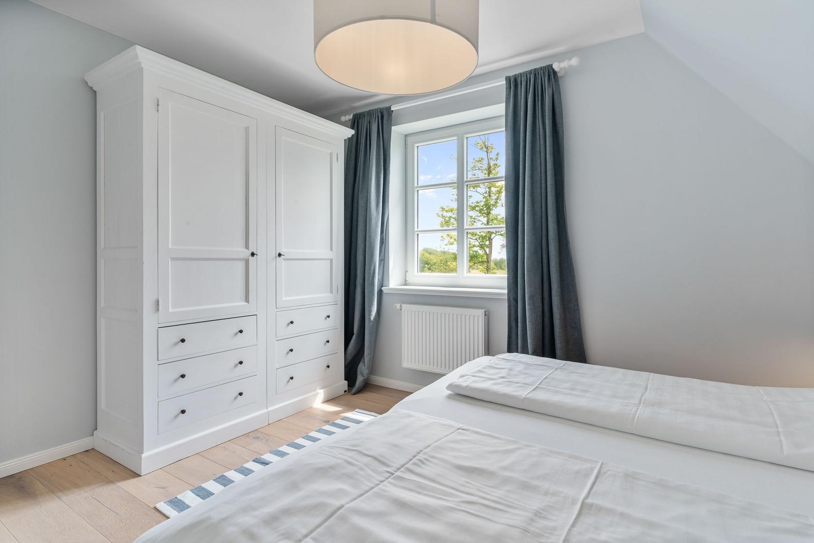 Schlafzimmer mit weißem Schrank, Bett und Fenster mit Blick auf Grün.