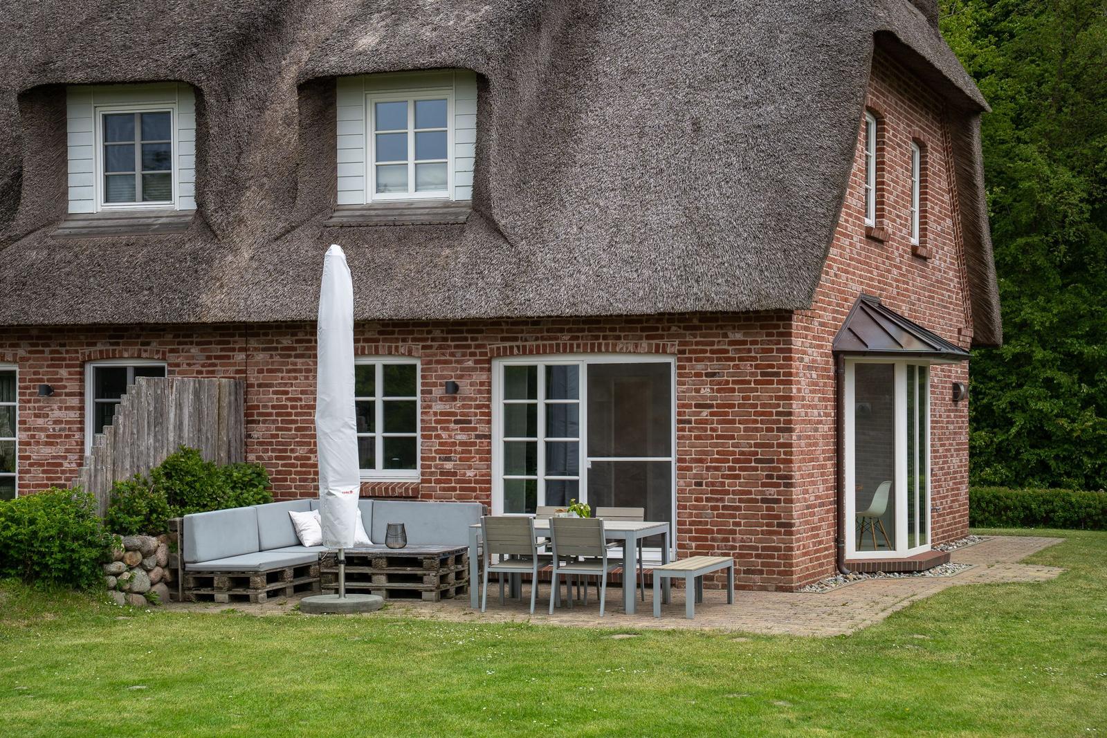Haus mit Strohdach, Terrasse mit Sitzgruppe und Tisch, Grasfläche und Bäume im Hintergrund.