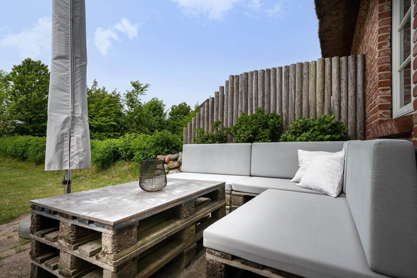 Terrasse mit grauem Sofa und Tisch aus Paletten. Hintergrund: Holzzaun und grüne Pflanzen.