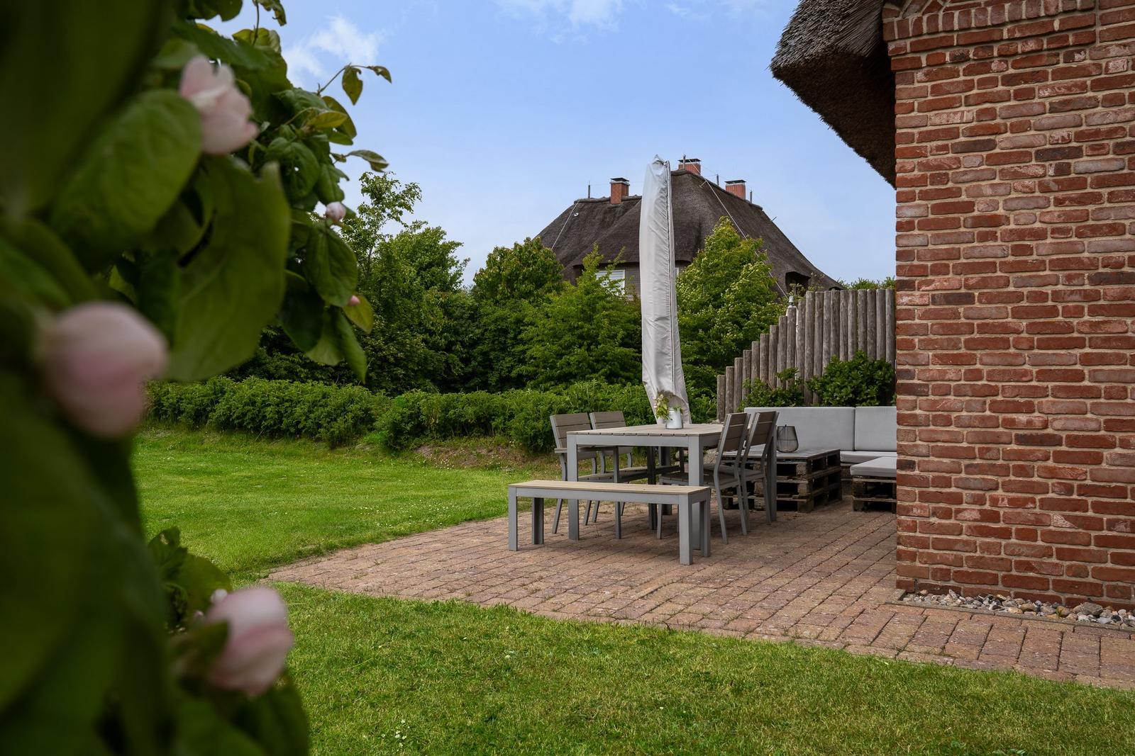 Terrasse mit Tisch, Stühlen und Sitzbank neben Backsteinmauer. Grasfläche und Rosenbusch im Vordergrund.