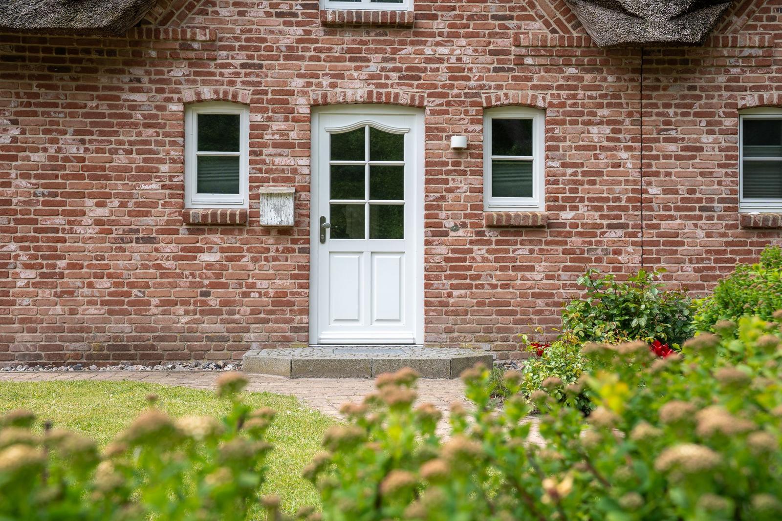 Haus mit rotem Ziegelsteinfassade, weißer Tür und Fenstern, gepflegter Garten mit Blumen.