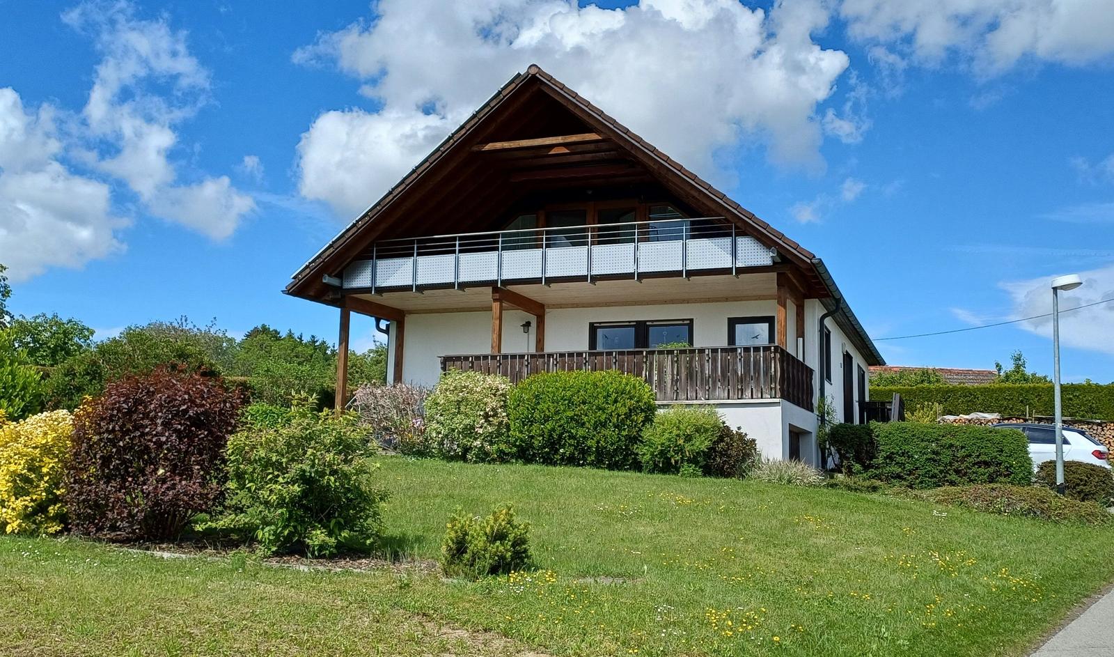 Ein zweistöckiges Haus mit Balkon, umgeben von Grünflächen und Bäumen unter blauem Himmel.
