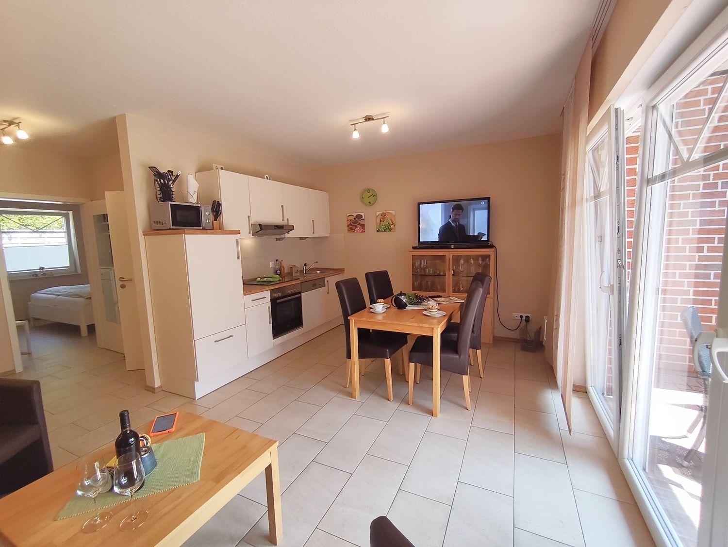 Wohnküche mit Essbereich, TV und Blick auf Balkon. Schlafzimmer im Hintergrund.