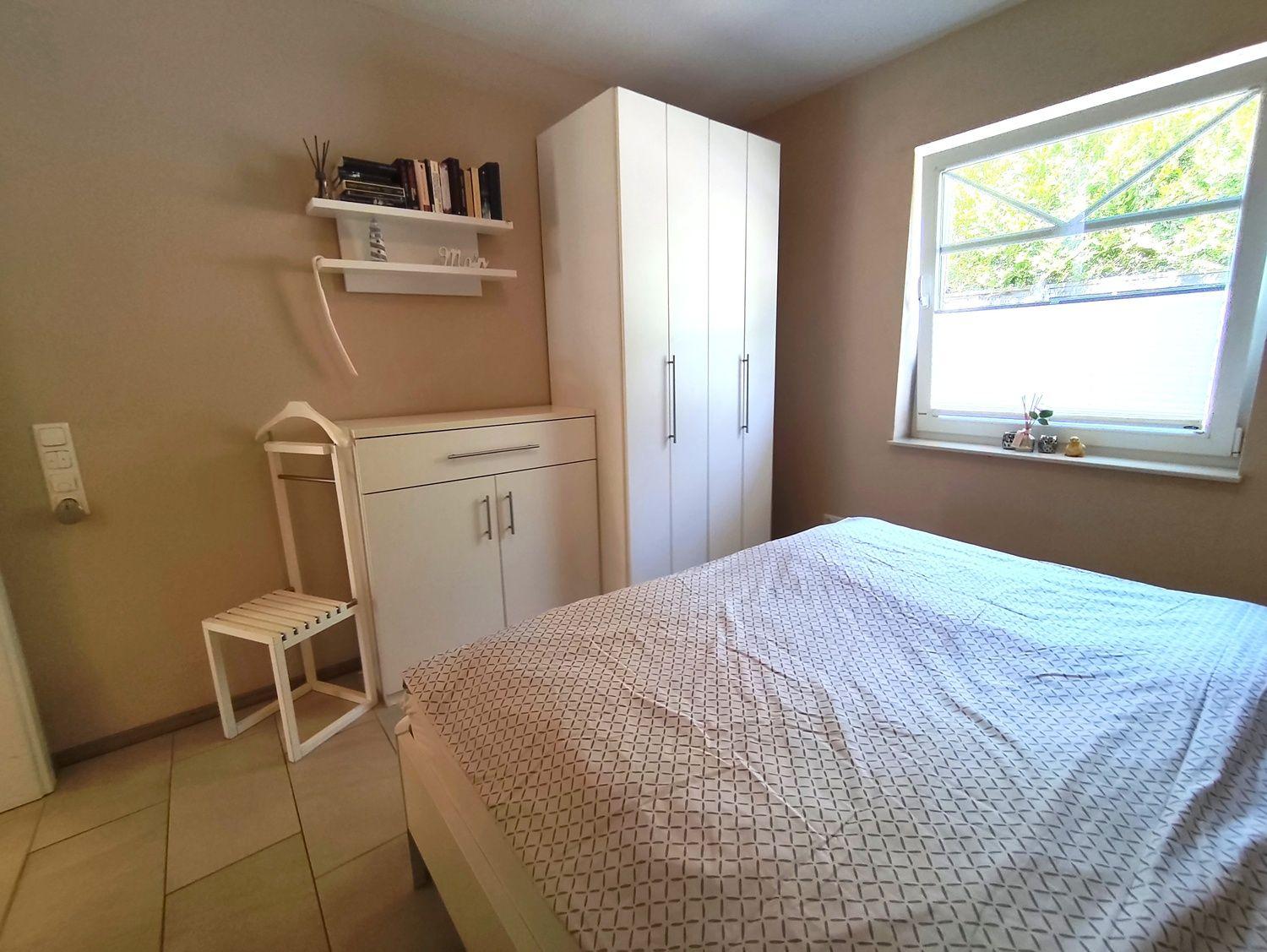 Schlafzimmer mit Bett, Schrank und Fenster. Auf dem Fensterbrett stehen kleine Dekorationsgegenstände.
