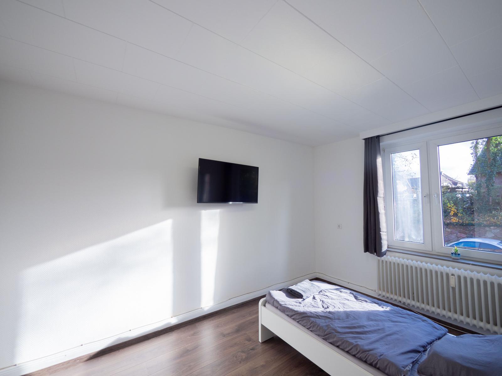 Schlafzimmer mit Bett, Fernseher und Fenster. Holzboden und weiße Wände.