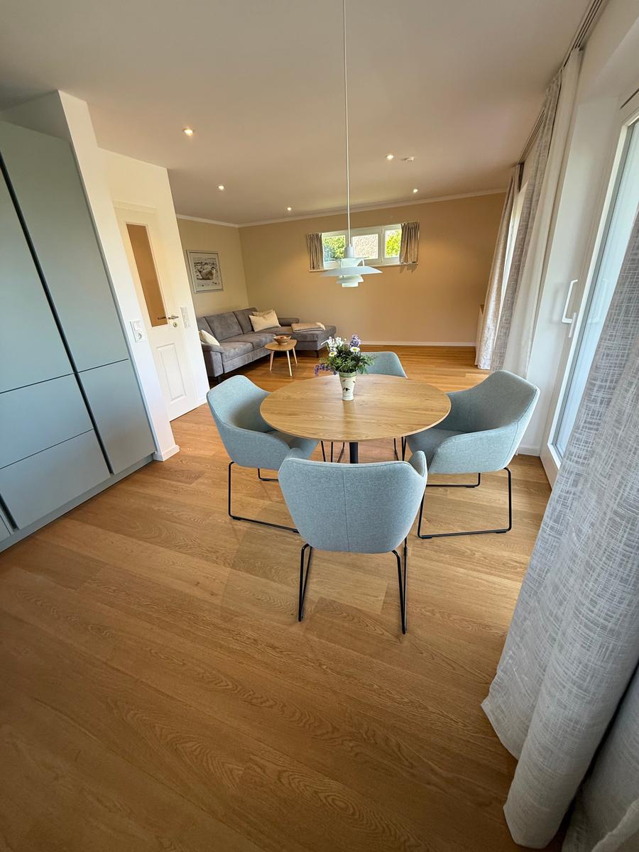 Essbereich mit runden Tisch und vier Stühlen. Im Hintergrund Wohnzimmer mit Sofa und Holzboden.