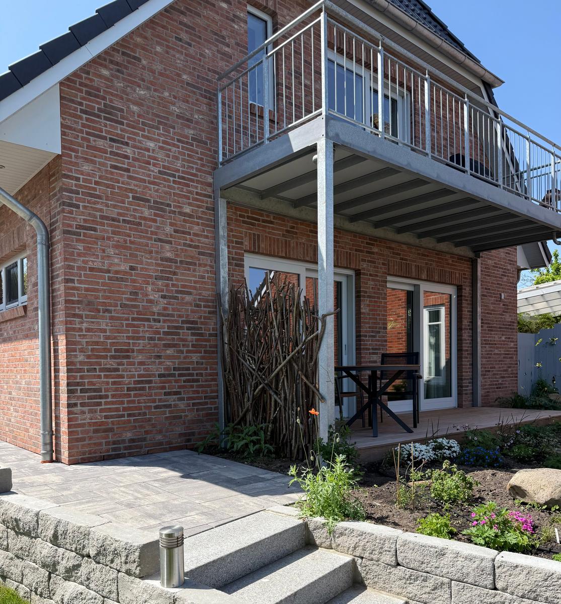 Rotes Backsteinhaus mit Balkon, Terrasse und Garten. Holztisch und Stühle stehen draußen.