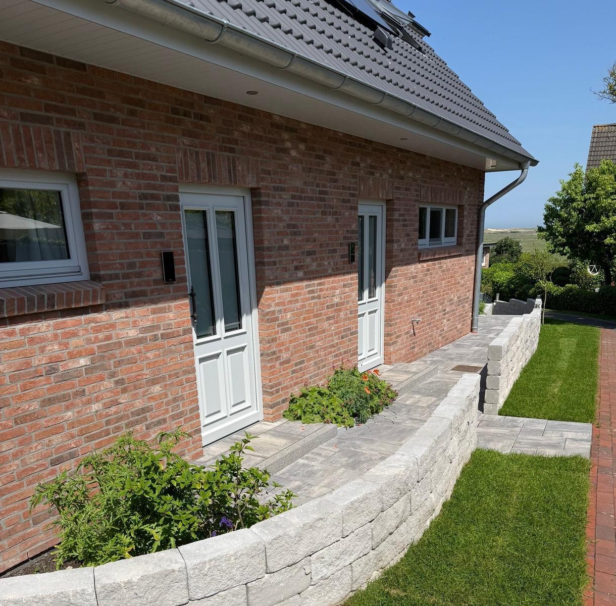 Rotes Backsteinhaus mit weißer Tür und gepflasterter Terrasse. Grünfläche und Steinmauer sichtbar.