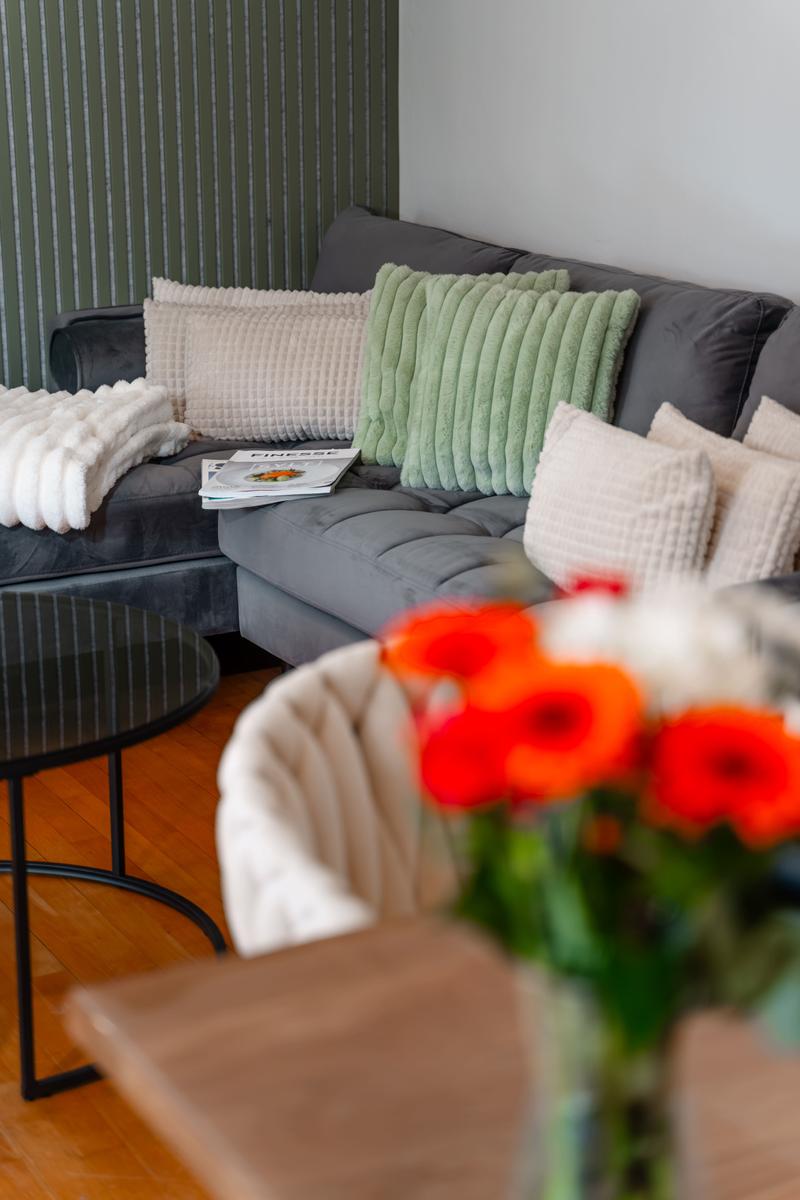 Ein grauer Sofa mit Kissen und einer Decke. Ein Tisch und Blumen sind im Vordergrund.