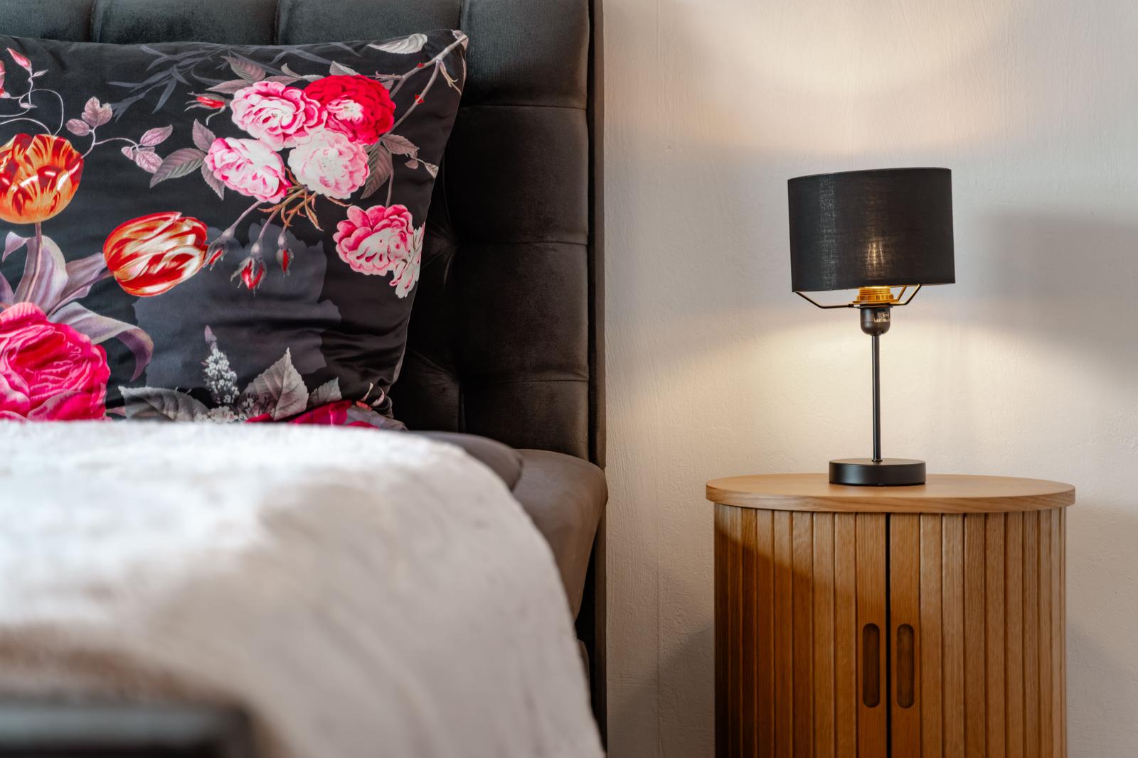 Schlafzimmer mit Bett, Kissen mit Blumenmuster und Nachttisch mit Lampe.