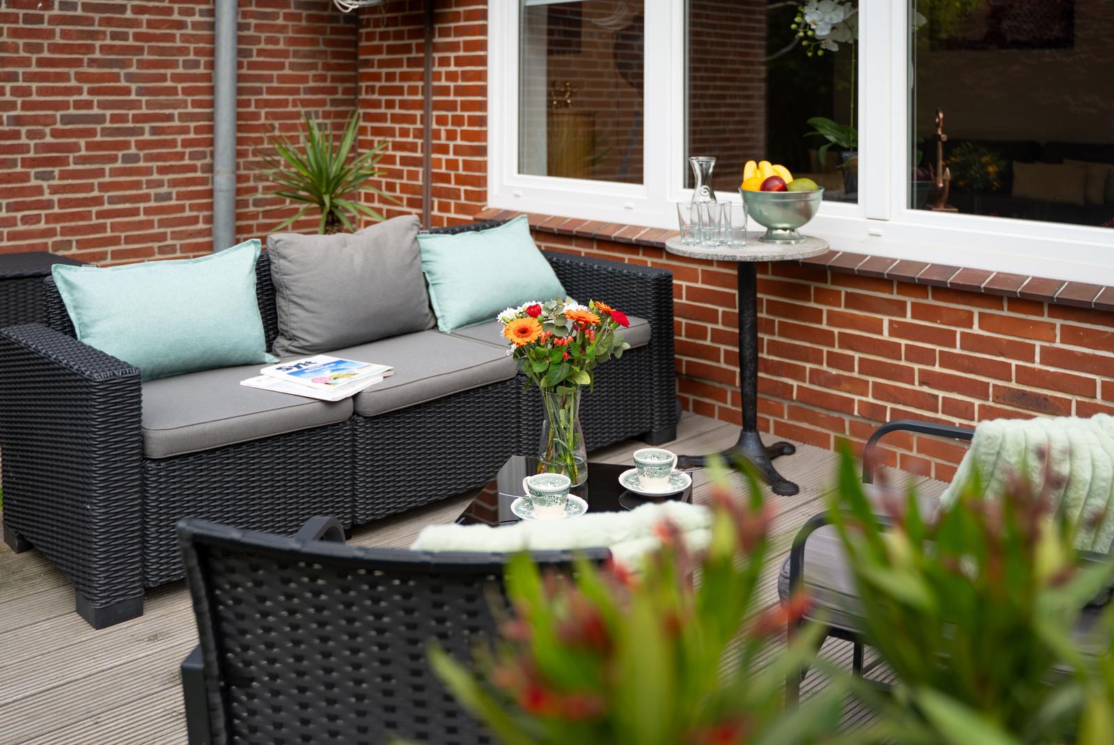 Terrasse mit Sitzgruppe, Blumen und Obstschale. Holzdielen und Backsteinmauer.