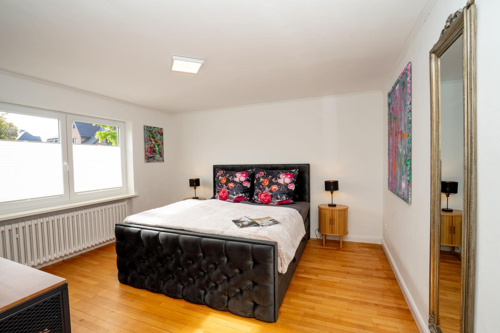 Schlafzimmer mit Bett, Nachttischen und Fenster. Holzboden und Wandbilder.