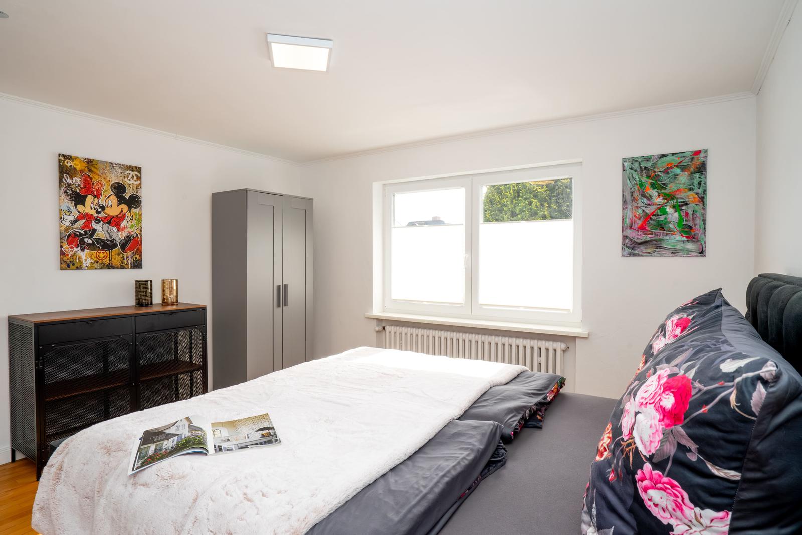 Schlafzimmer mit Bett, Kleiderschrank und Fenster. Wandbilder und Zeitschrift auf Bett.