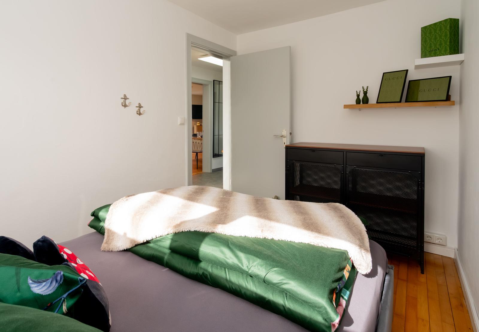 Schlafzimmer mit Bett, Schrank und Wandregal. Tür führt in Wohnbereich.