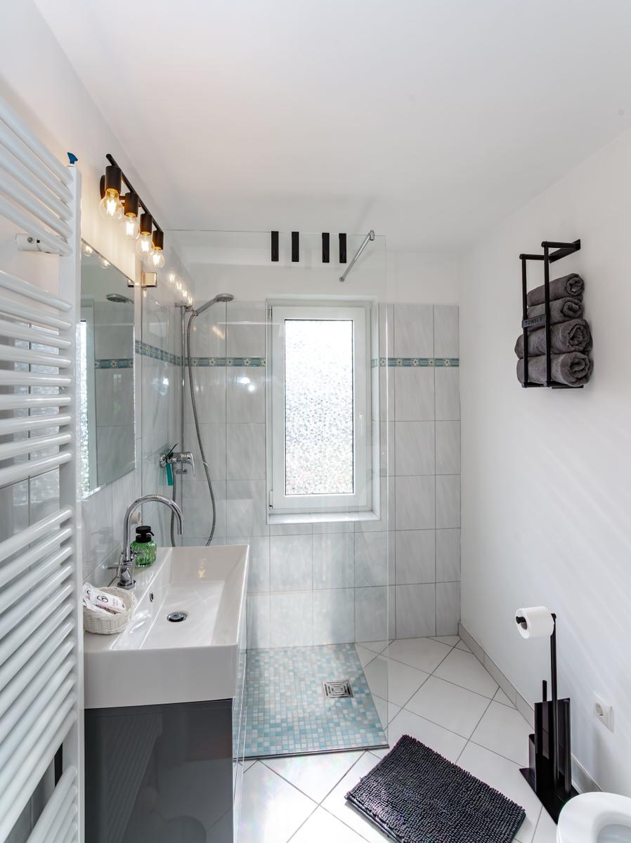 Badezimmer mit Dusche, Waschbecken und Toilette. Fenster, Handtuchhalter und Heizung sichtbar.