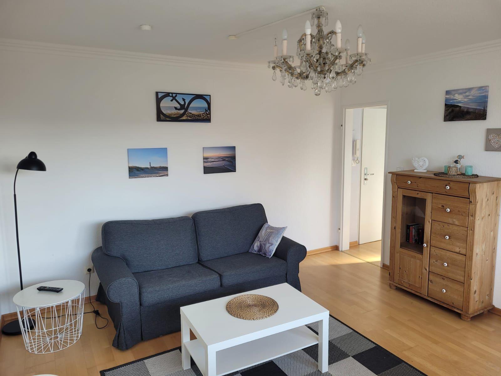 Wohnzimmer mit grauem Sofa, weißem Couchtisch und Holzschrank. An den Wänden hängen Bilder.