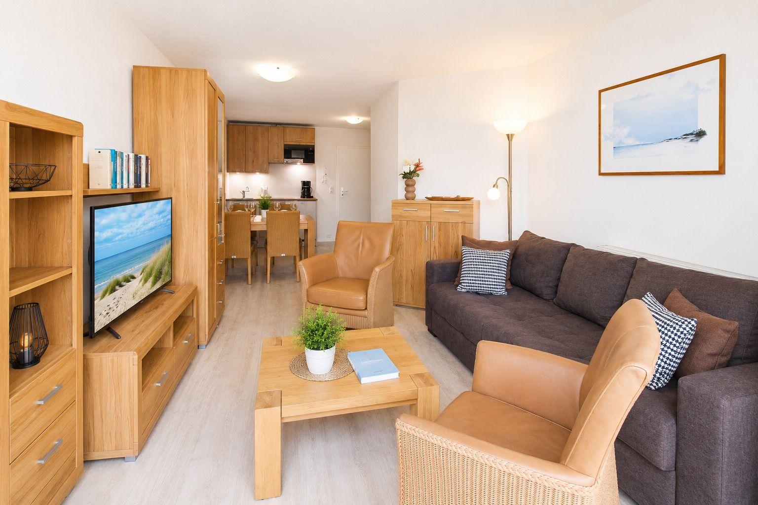 Wohnzimmer mit Sofa, Sessel und TV. Küche und Essbereich im Hintergrund.