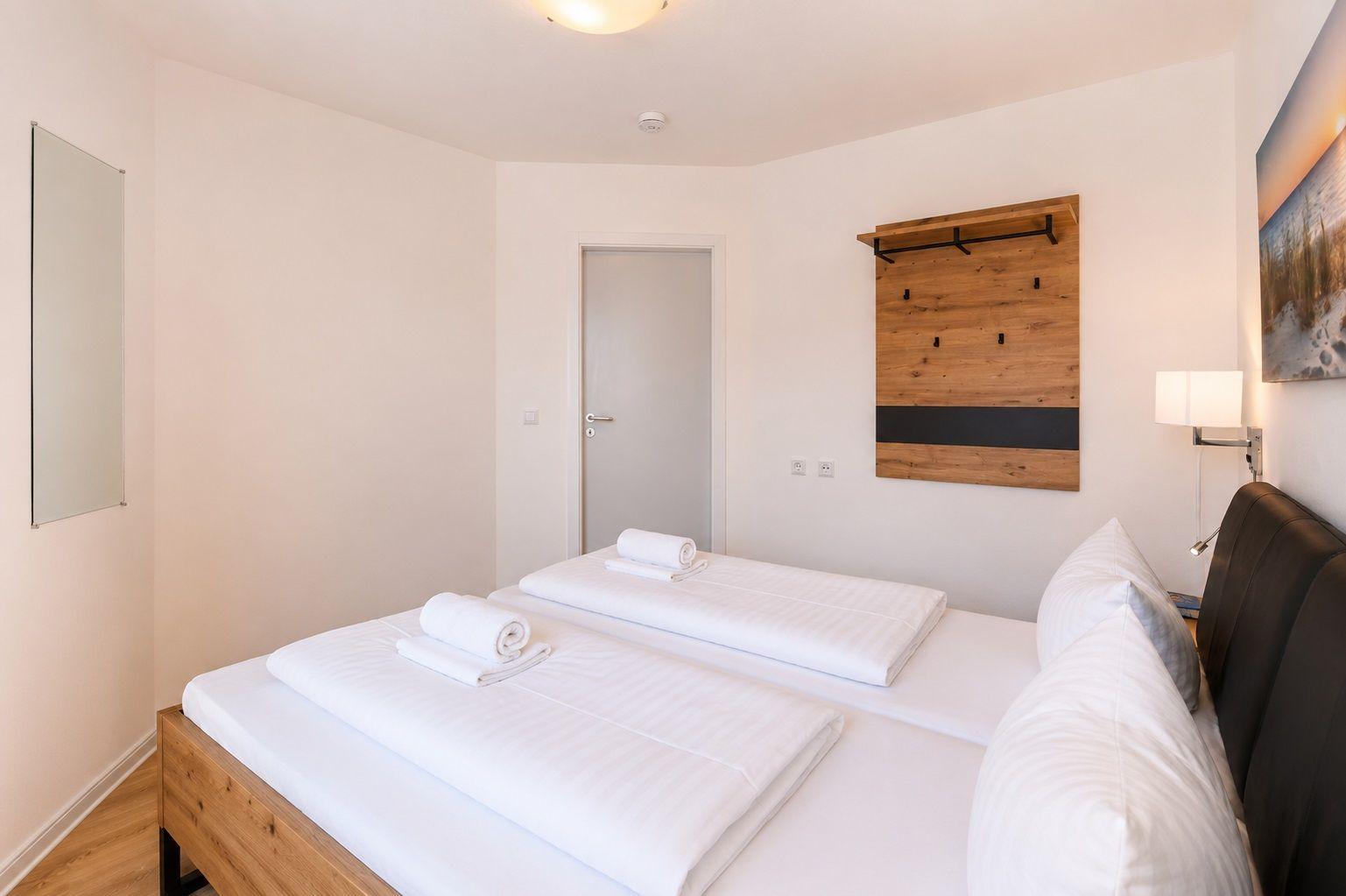 Schlafzimmer mit Doppelbett, Holzgestell, Wandregal und Spiegel an der Wand.