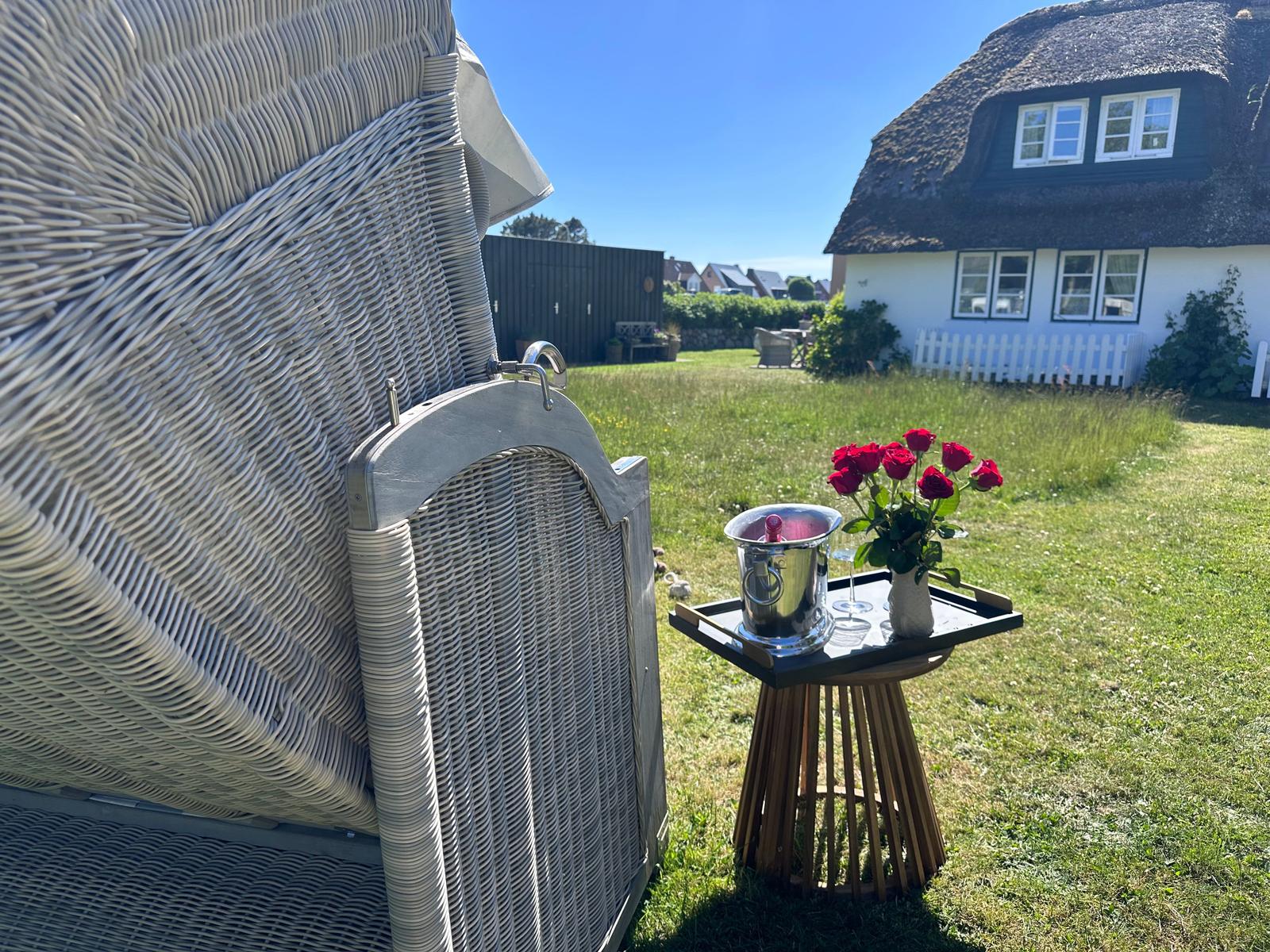 Wickerliege mit Tablett, roten Rosen und Eiswasser im Garten vor einem Haus mit Strohdach.