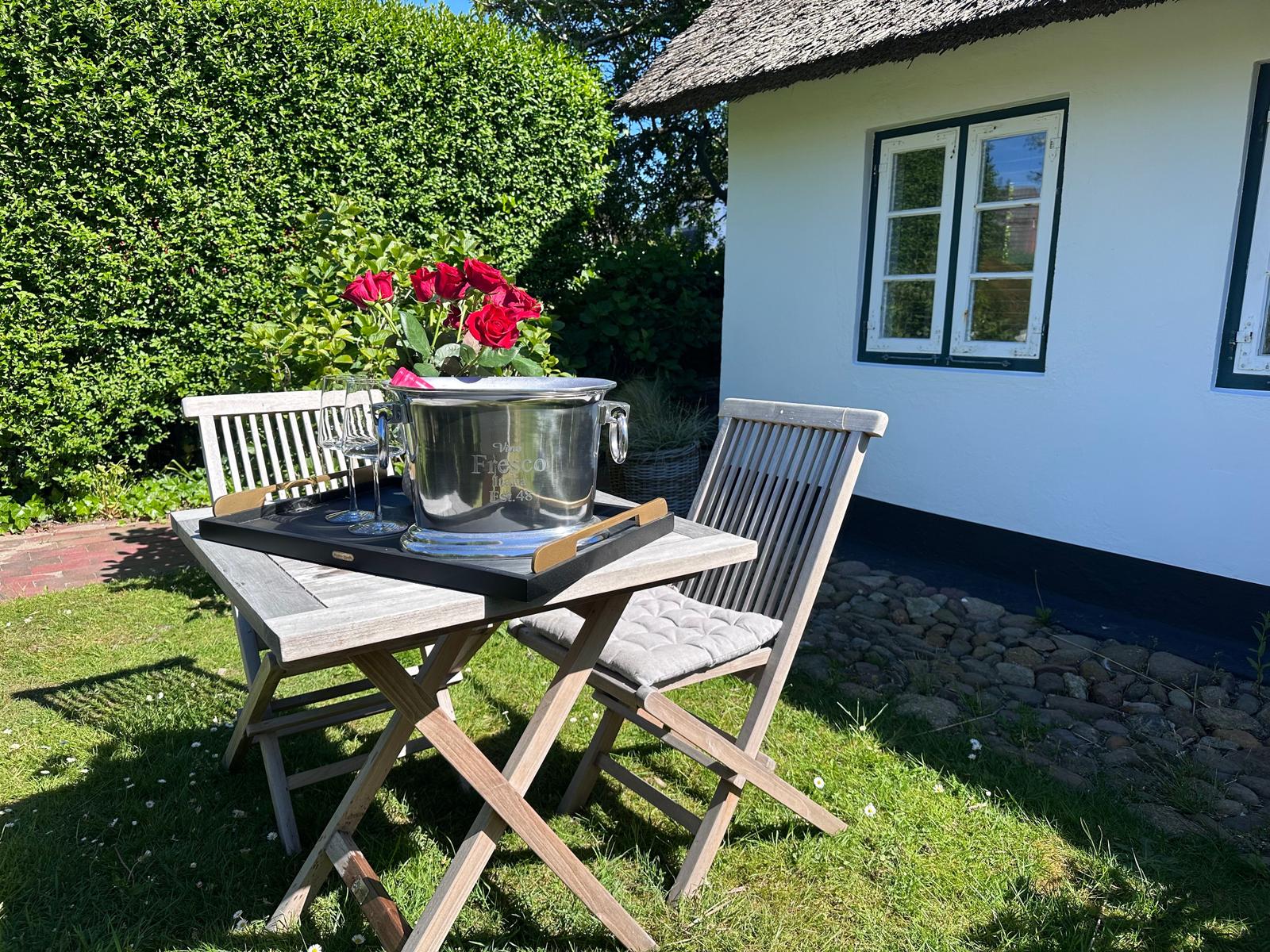Holztisch mit Stühlen, Kühler und Rosen im Garten vor einem Haus.