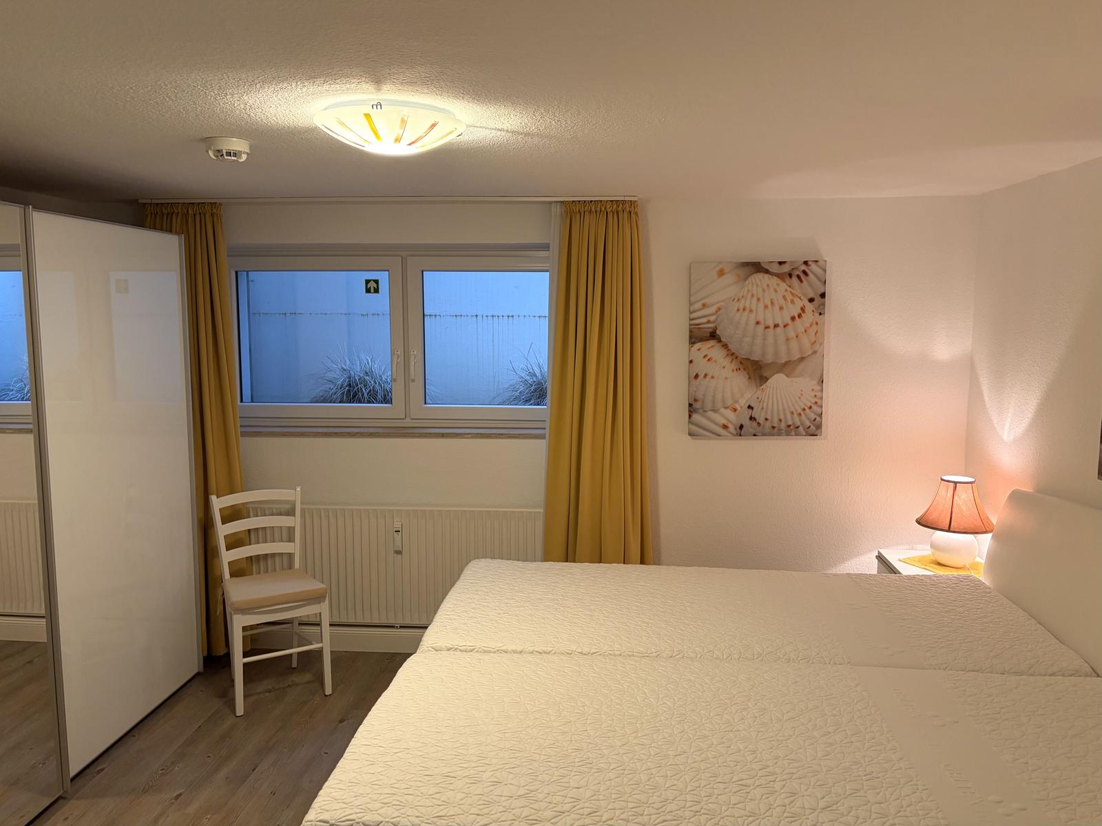 Schlafzimmer mit Bett, Nachttisch, Lampe und Fenster mit gelben Vorhängen.