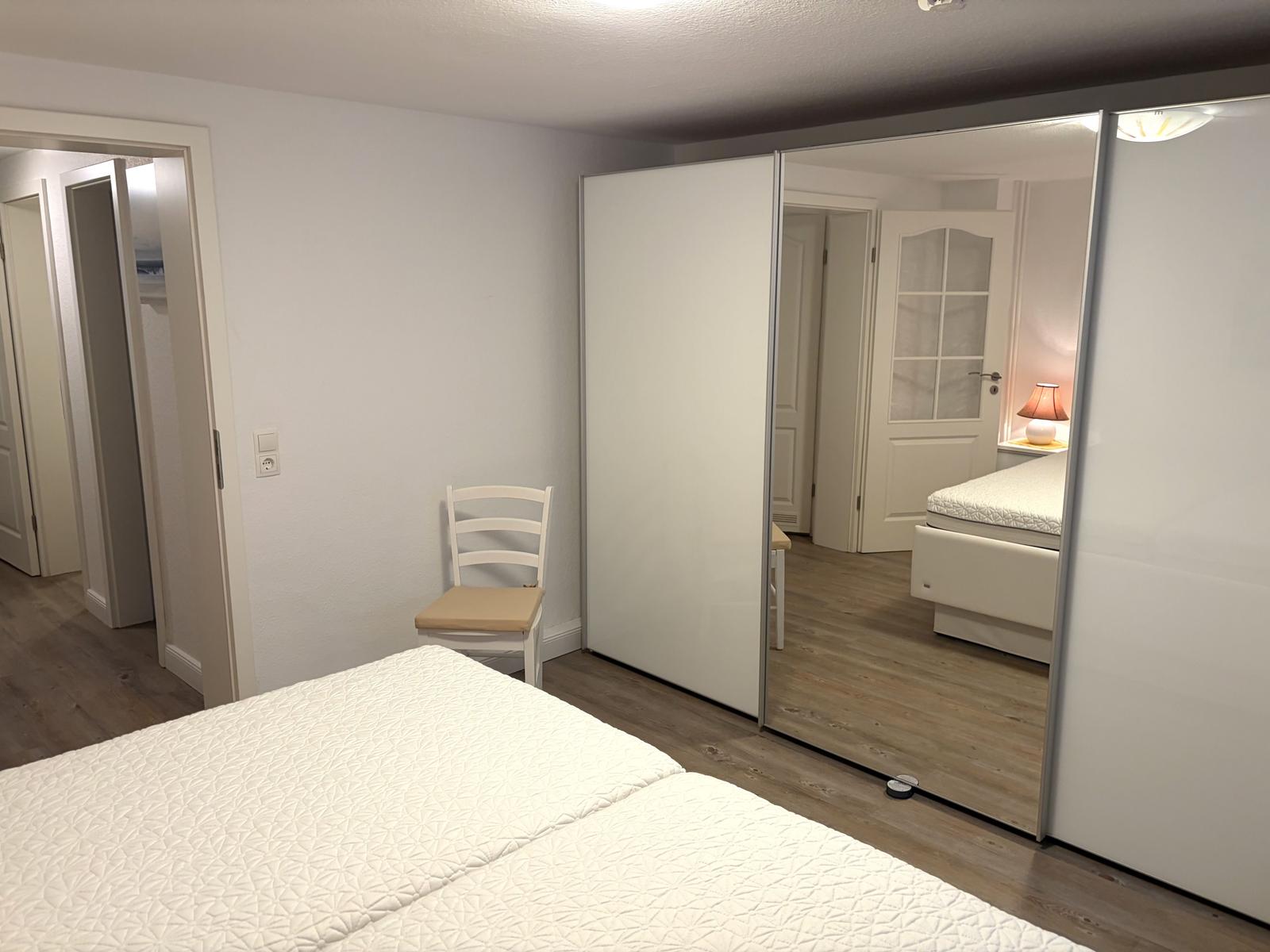Schlafzimmer mit Bett, Stuhl und Schiebetüren. Spiegel reflektiert weitere Räume.