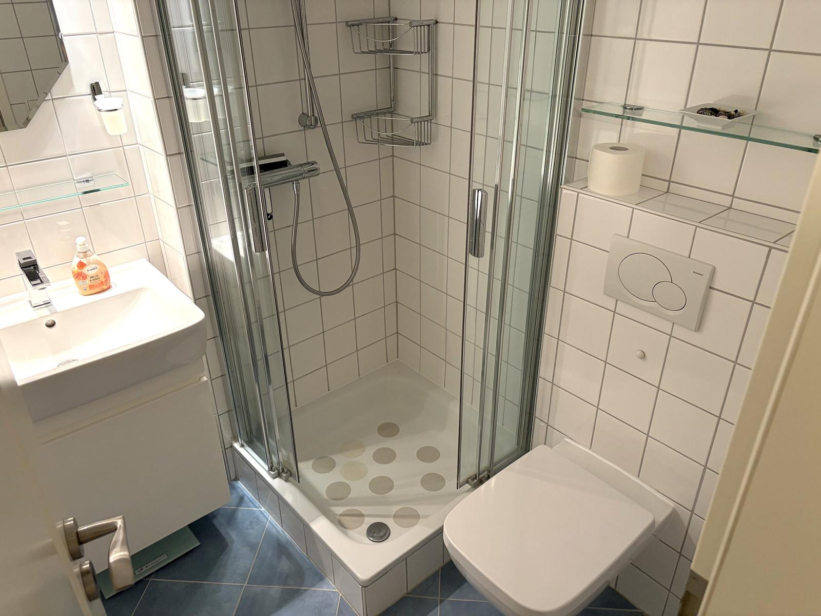 Badezimmer mit Dusche, Waschbecken und WC. Fliesenboden und weiße Wände.