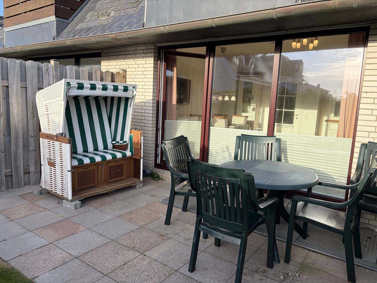 Terrasse mit Strandkorb und Tisch mit Stühlen vor Haus mit Glasfront.