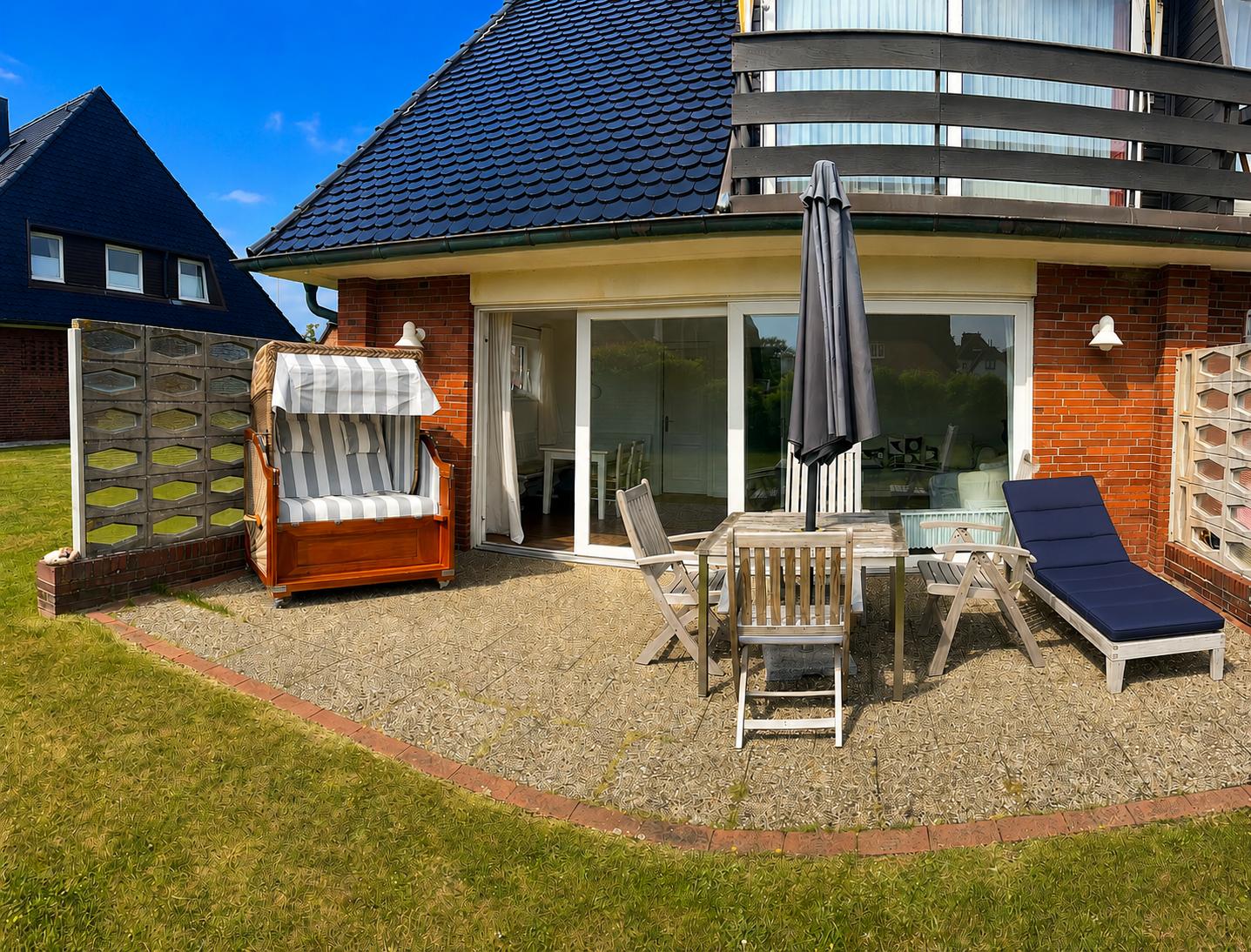 Terrasse mit Strandkorb, Tisch, Stühlen und Liege. Haus mit Glasfront und Balkon.