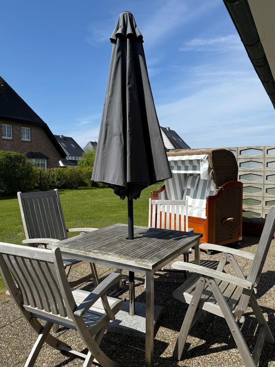 Terrasse mit Tisch, vier Stühlen und geschlossenem Sonnenschirm. Im Hintergrund Strandkörbe und Grünfläche.