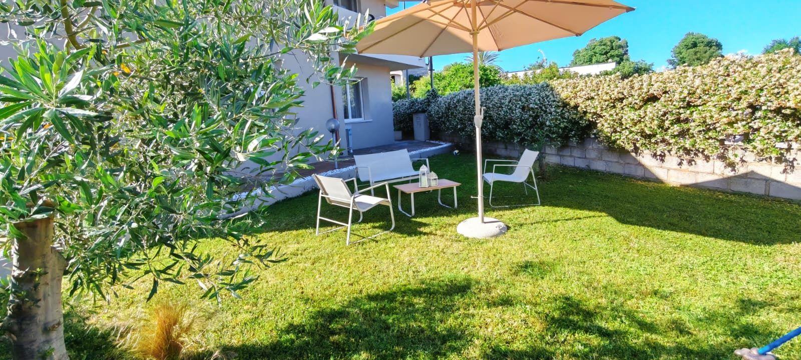 Garten mit Sonnenschirm, drei Stühlen und Tisch. Grasfläche, Baum und Hecke im Hintergrund.