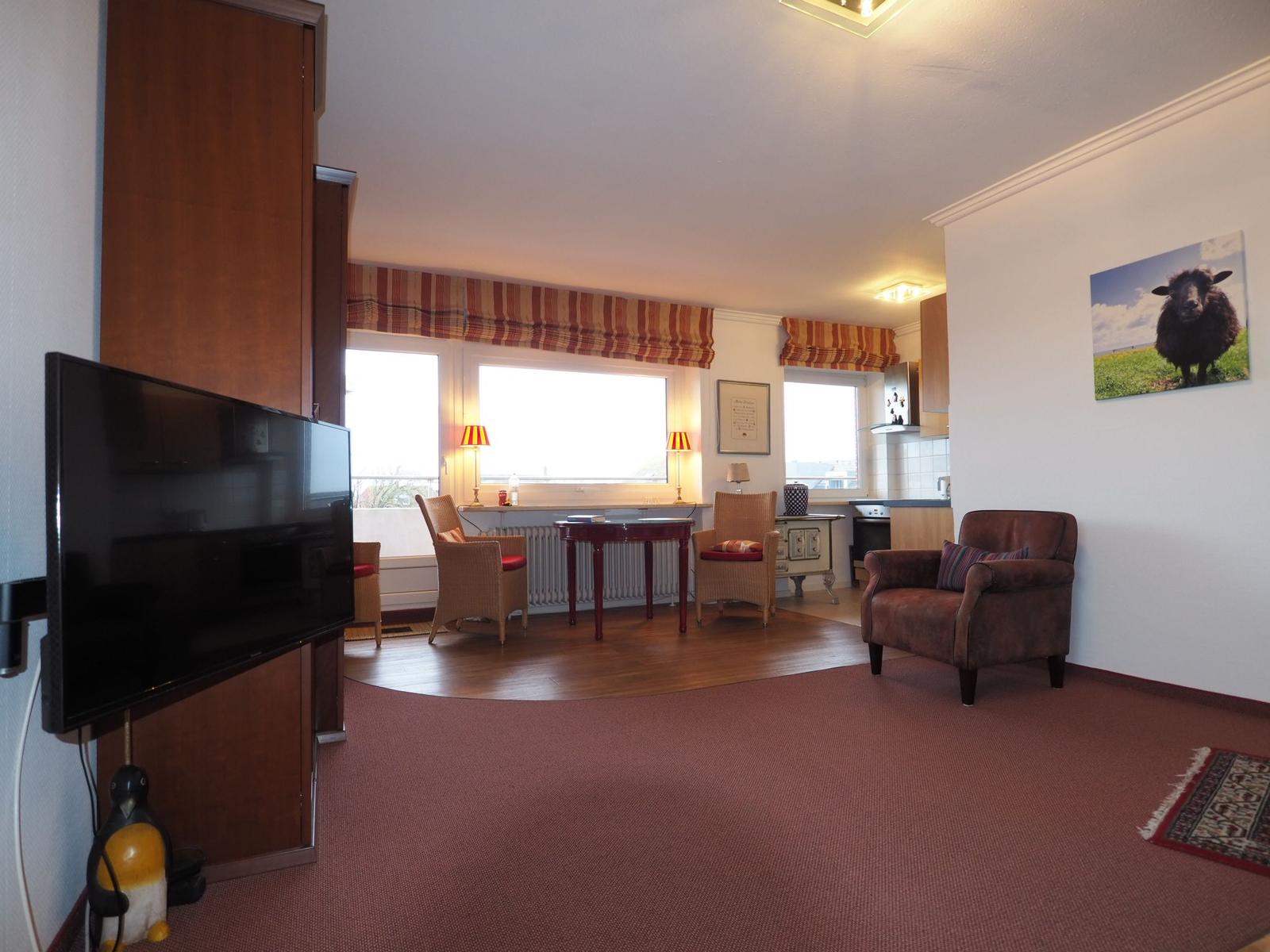 Wohnraum mit Fernseher, Sitzgelegenheiten und Fenstern mit Rollos.
