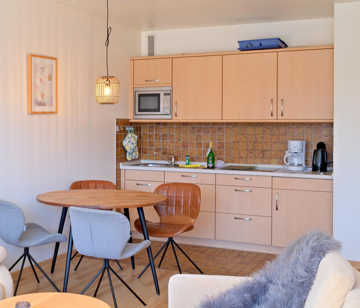 Küche mit Holzschränken, Esstisch und Stühlen. Hintergrund: Fliesenwand und Mikrowelle.